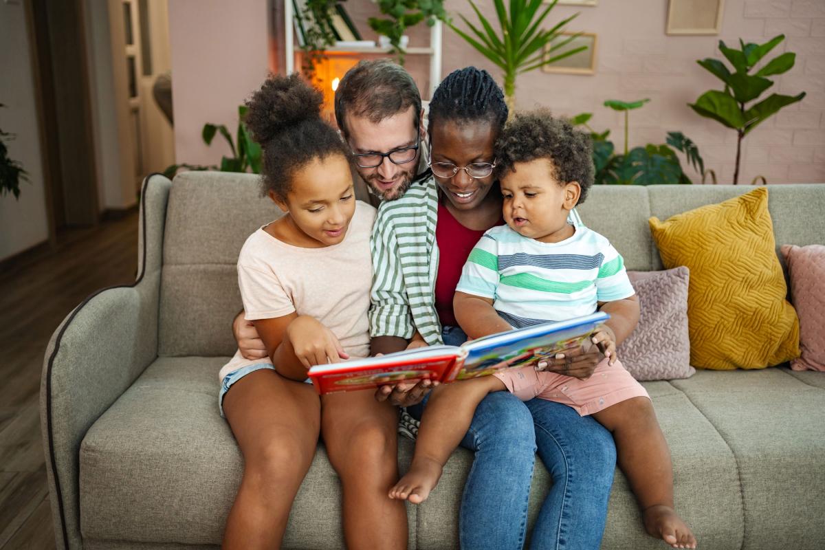 a family with an adult male, adult female, and 2 children reading a story on a couch. 