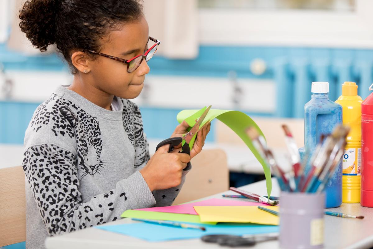 young black girl with glasses cuts a piece of green construction paper at a desk