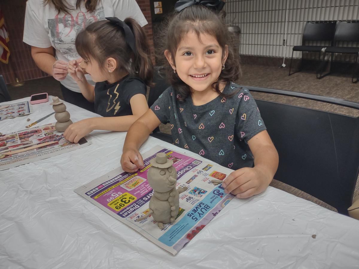 Small child with dark hair smiling and working on a clay piece at a table.