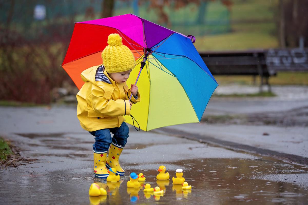 toddler holding a rainbow umbrella plays near a puddle that has toy rubber ducks in it