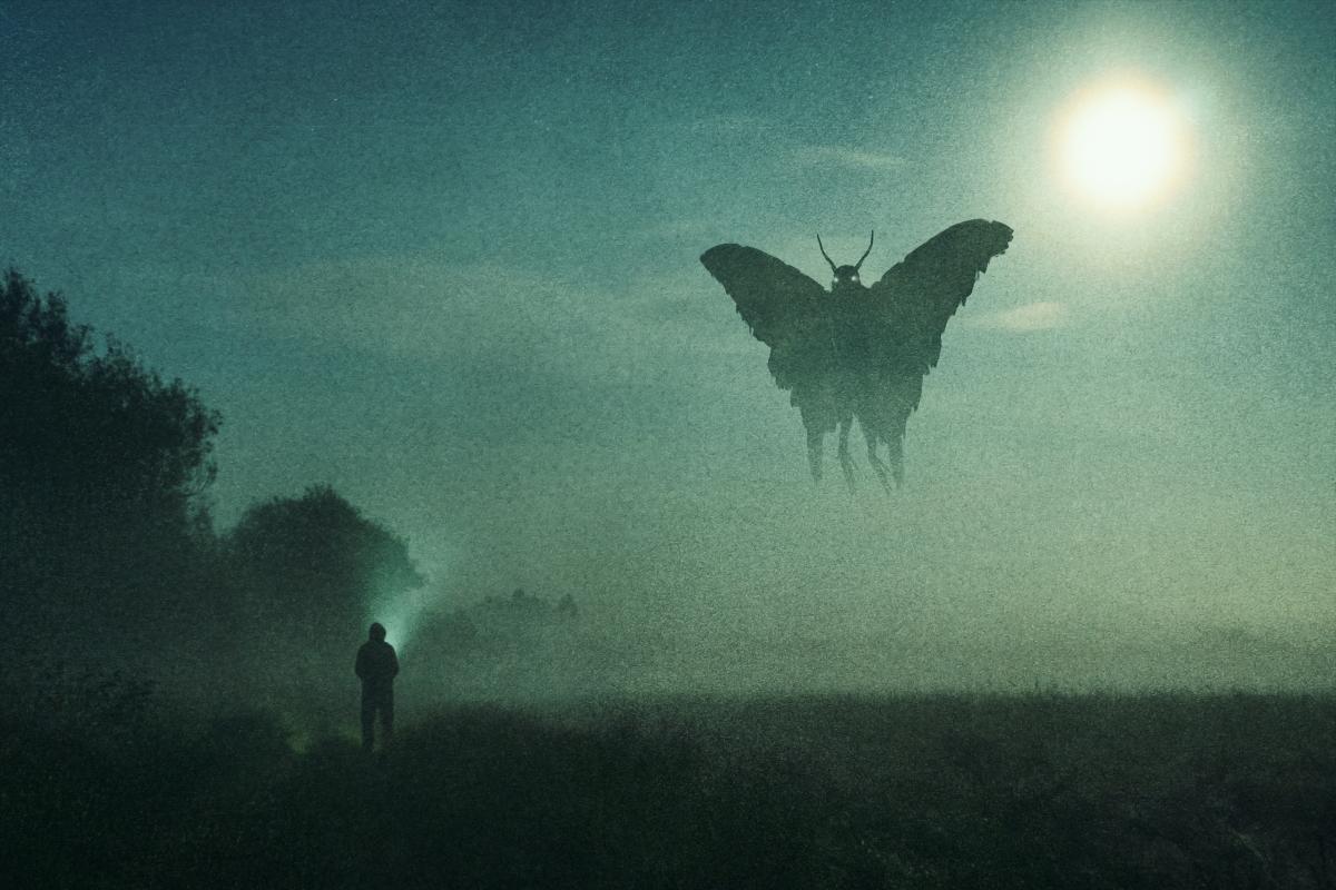 An open foggy field shows a man standing as a Mothman hovers over him in the moonlight. 