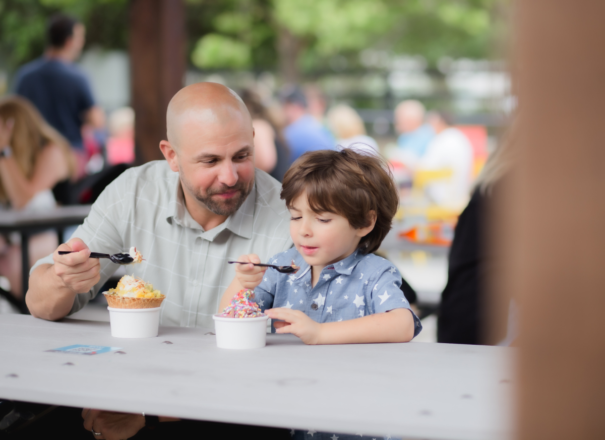 A man and a young boy sit at a table and eat ice cream out of cups. 