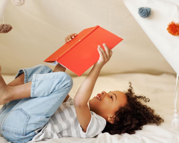 young black girl laying on her back in a soft play tent reading a red book 