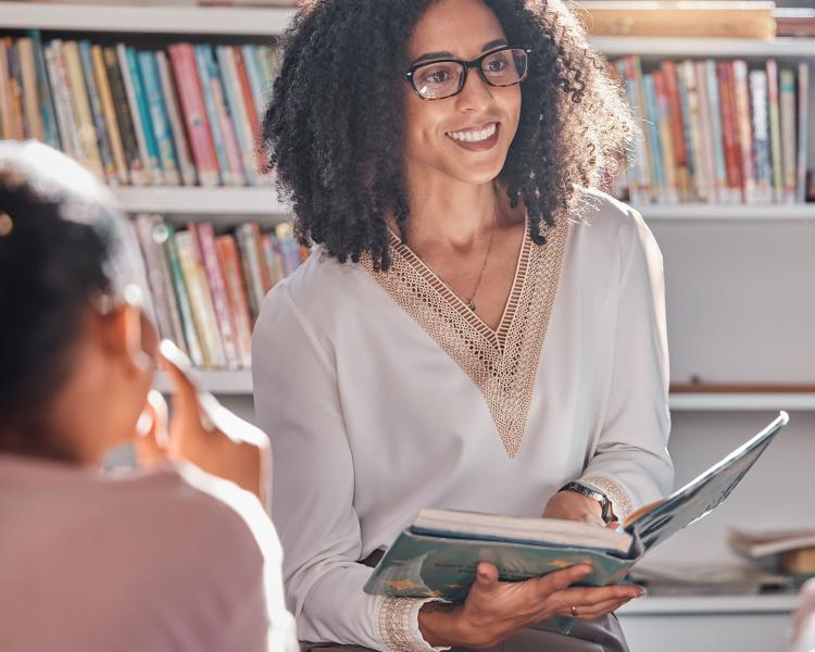 black woman with sunlight behind her sits on floor of library reading to children